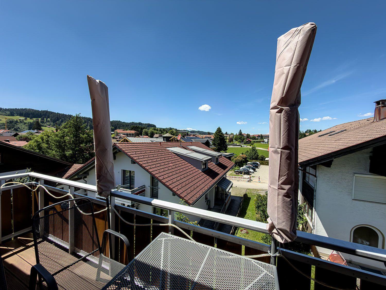 Balcony with table and chairs, view of rooftops and green hills under blue sky.
