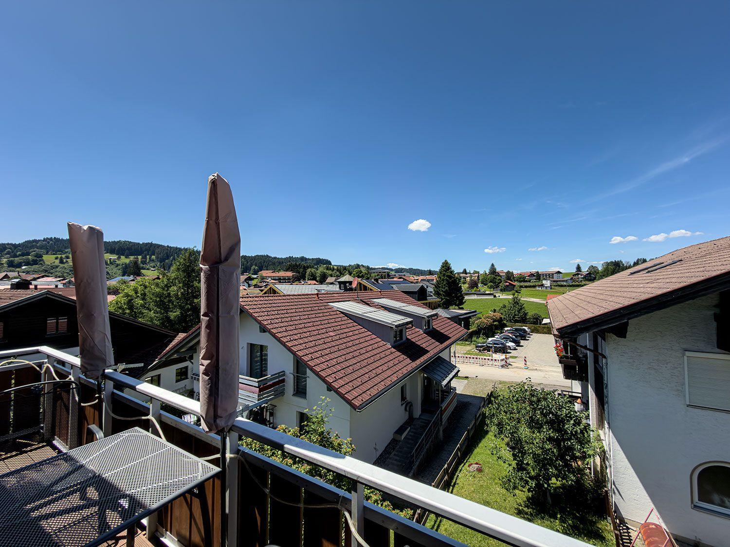 Terrace with table and closed umbrellas. View of roofs, green areas, and mountains under blue sky.