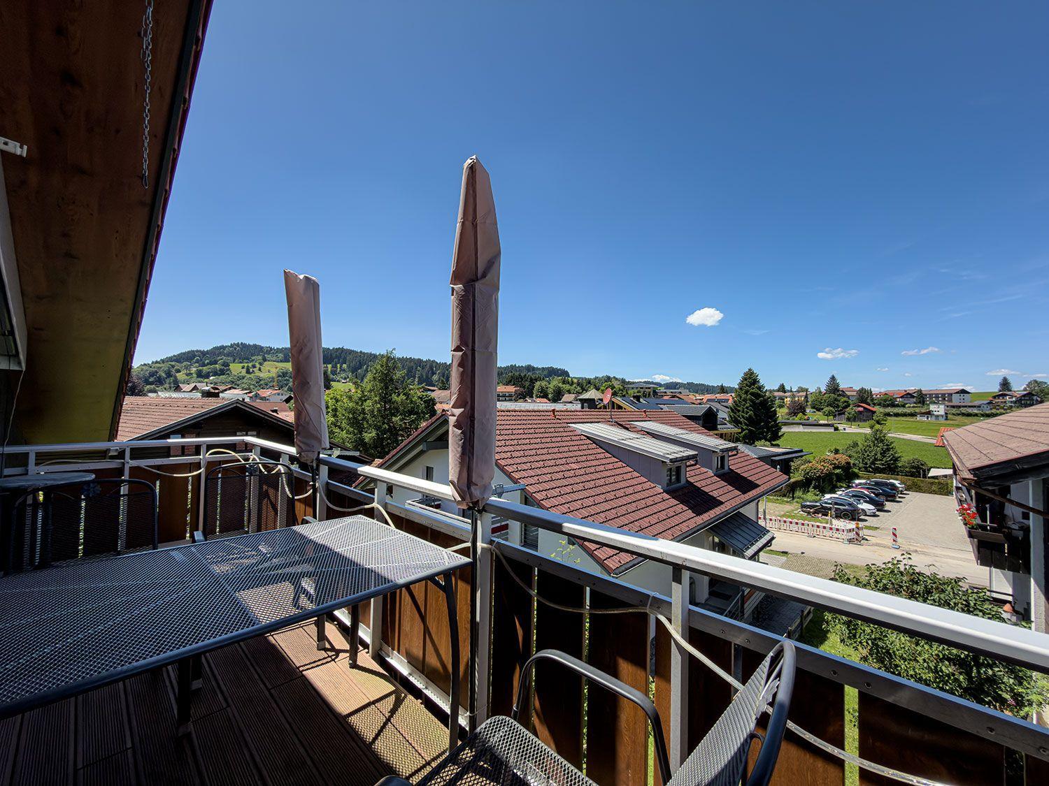 Terrace with table and chairs, view of rooftops and green hills under blue sky.