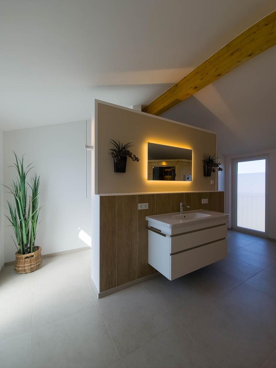 Badezimmer mit Waschbecken, Spiegel und Pflanzen. Holzdeckenbalken und große Fenster.