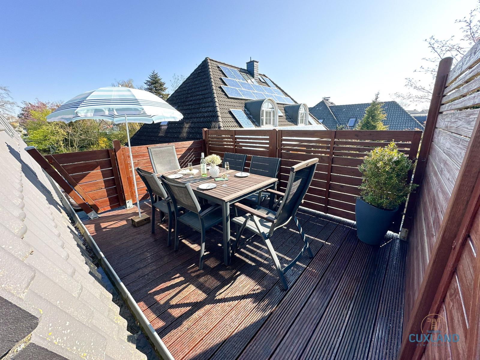 Terrasse mit Tisch, Stühlen und Sonnenschirm. Holzgeländer und Pflanze sind sichtbar.