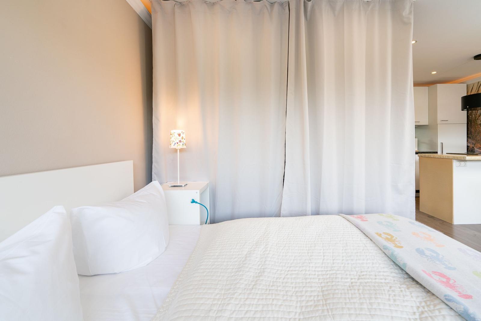 Schlafzimmer mit Bett, Nachttisch und Vorhang. Küche im Hintergrund sichtbar.