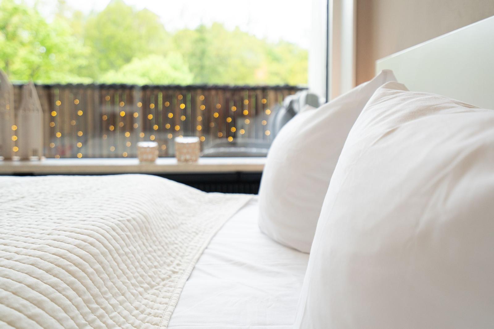 Ein Bett mit weißen Kissen und einer gesteppten Decke vor einem Fenster mit Lichterketten.