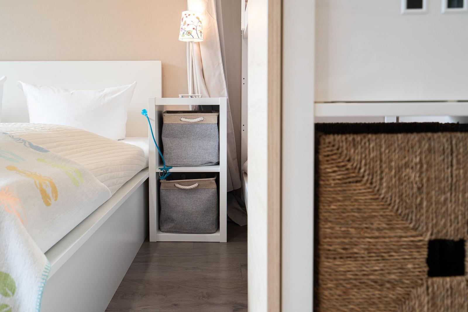 Schlafzimmer mit Bett, Nachttisch und Körben. Lampe und Vorhang sind sichtbar.