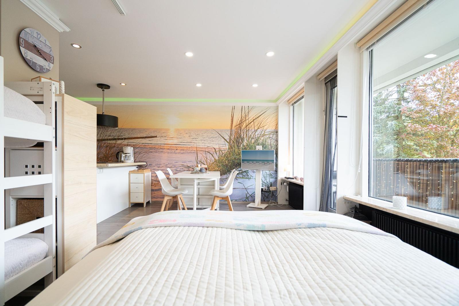 Schlafzimmer mit Blick auf Balkon, Küchenbereich und Wandbild mit Strandmotiv.