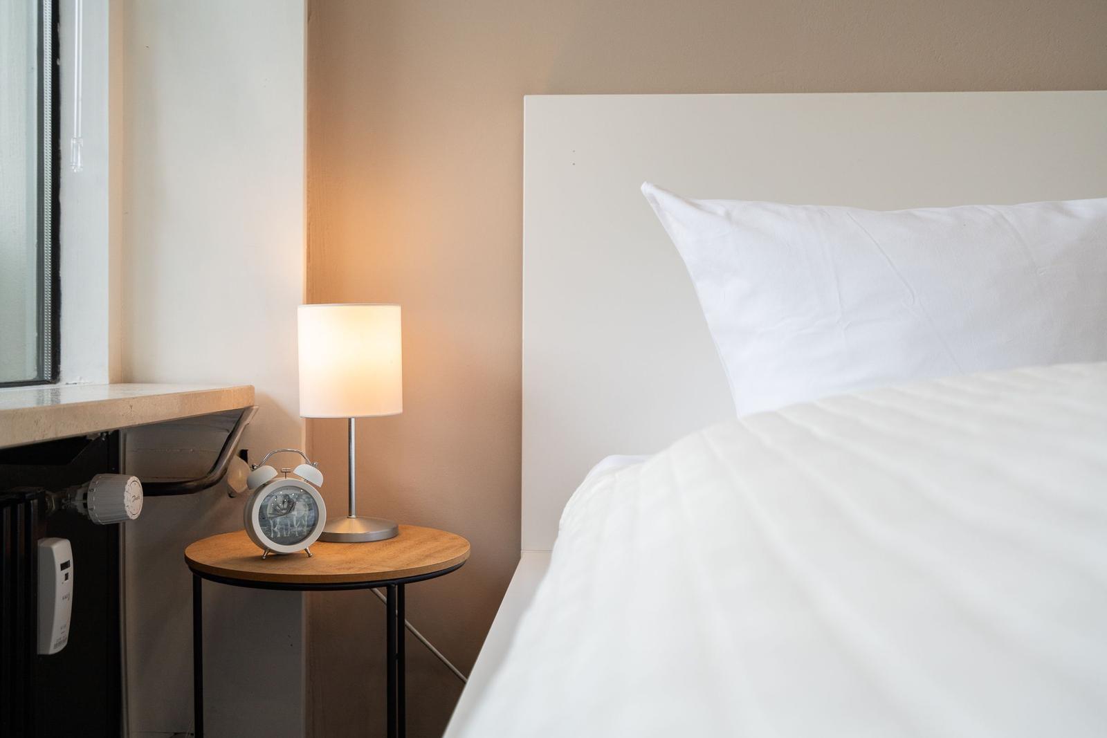 Ein Bett mit weißer Bettwäsche, Nachttisch mit Lampe und Wecker.