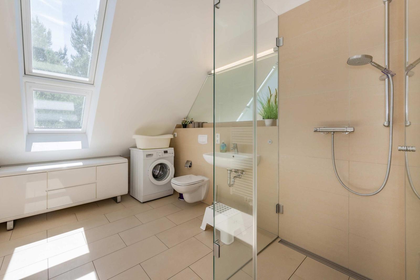 Badezimmer mit Dusche, Waschmaschine und Fenster. Fliesenboden und Spiegel.