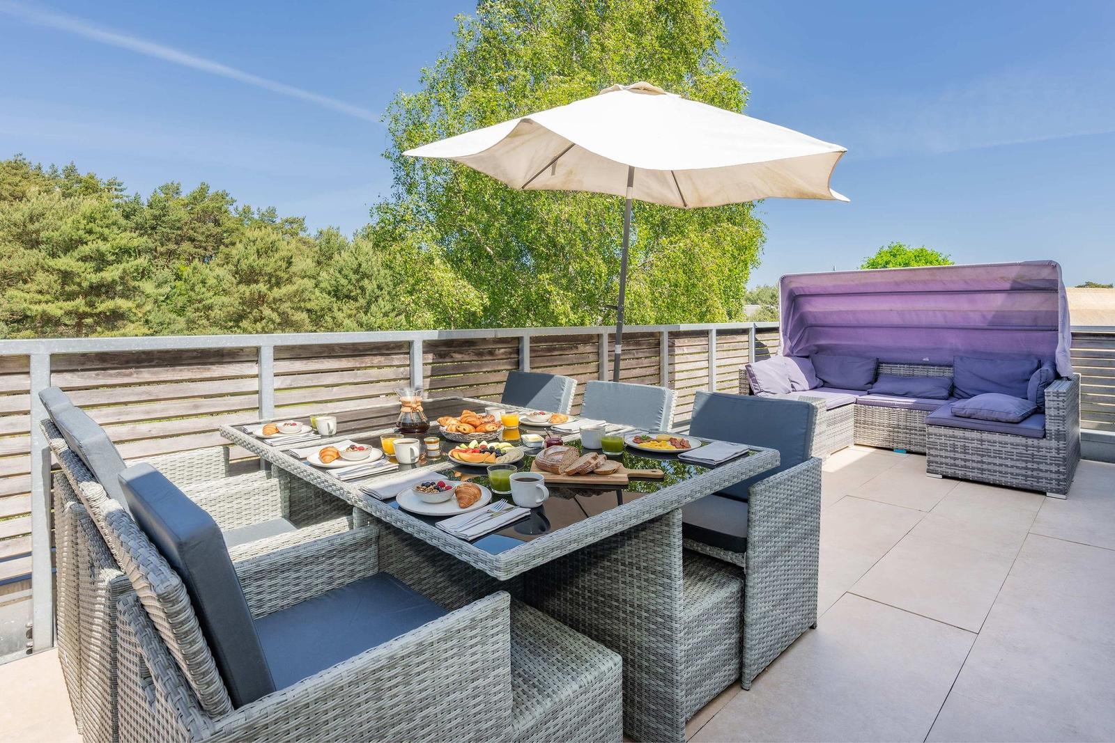 Terrasse mit Esstisch, Sitzgruppe und Sonnenschutz. Hintergrund: Wald und klarer Himmel.