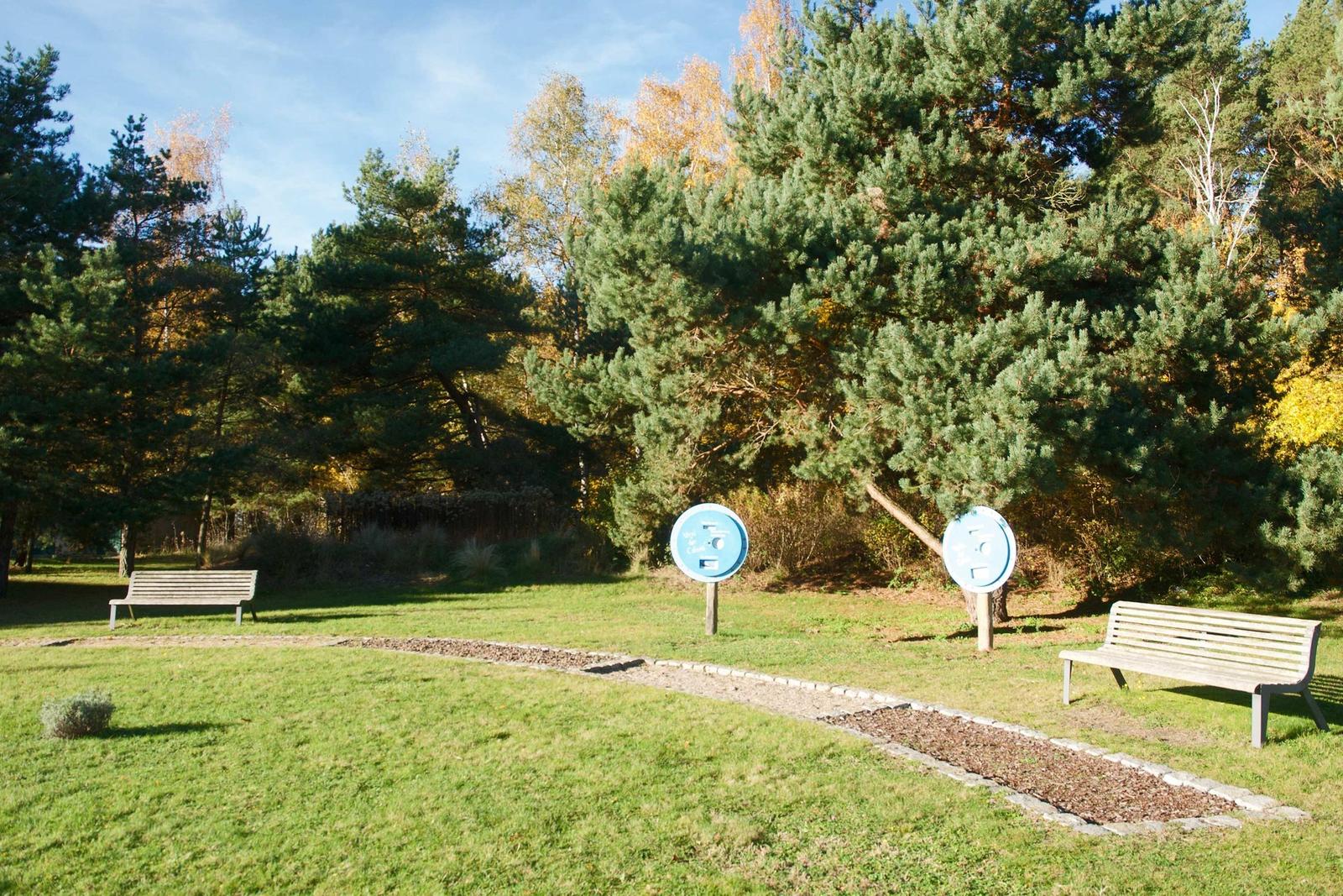 Grüner Garten mit Bänken und Bäumen. Zwei blaue Schilder stehen im Gras.