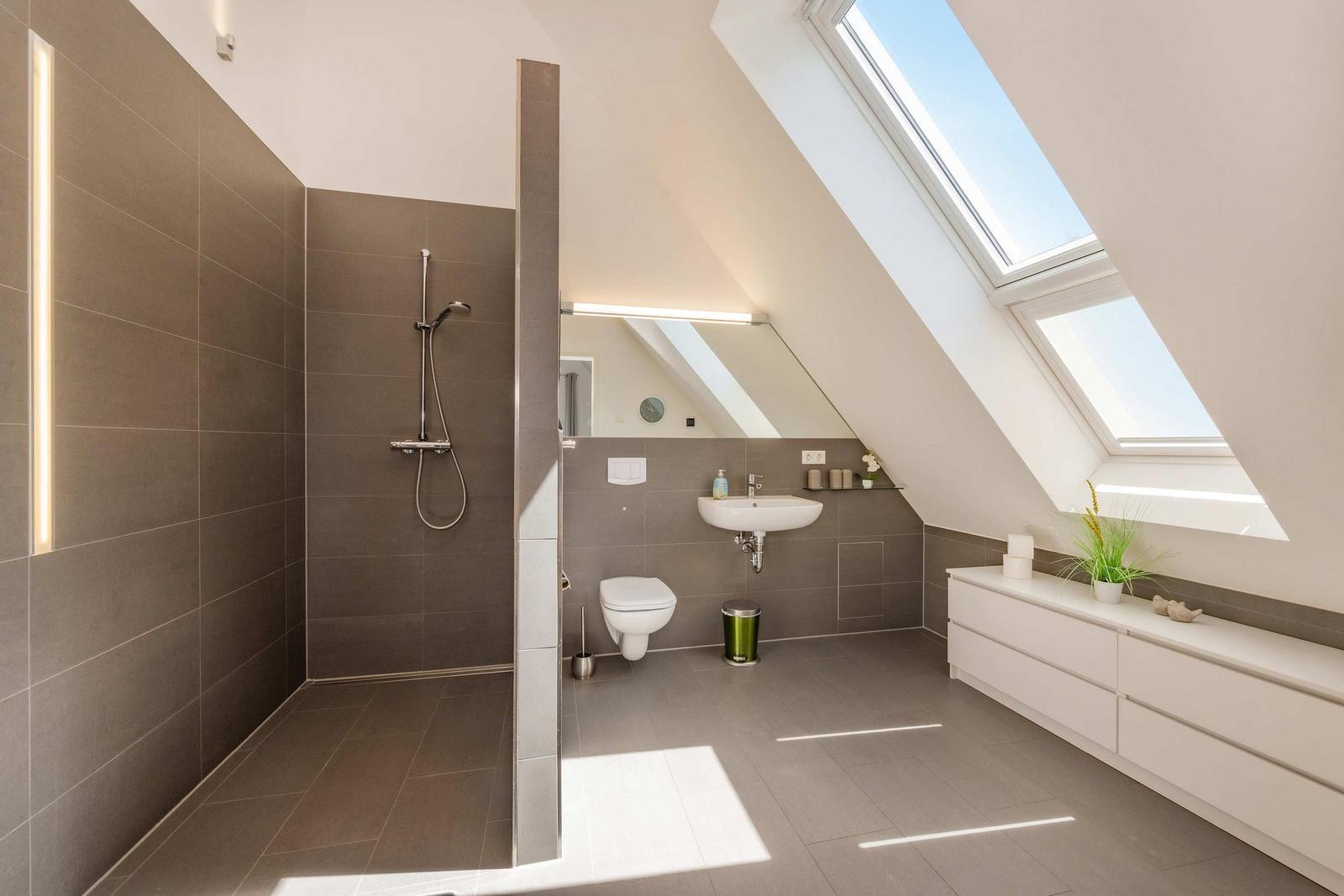 Badezimmer mit Dusche, Waschbecken und Toilette unter Dachschrägen mit Fenstern.