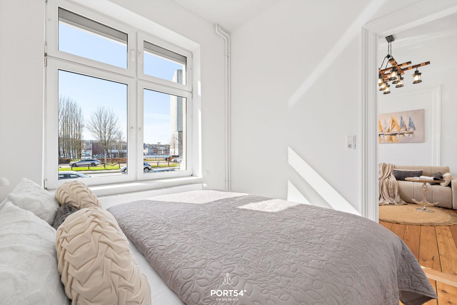 Schlafzimmer mit großem Fenster und Blick auf die Straße. Tür führt in Wohnbereich mit Sofa.