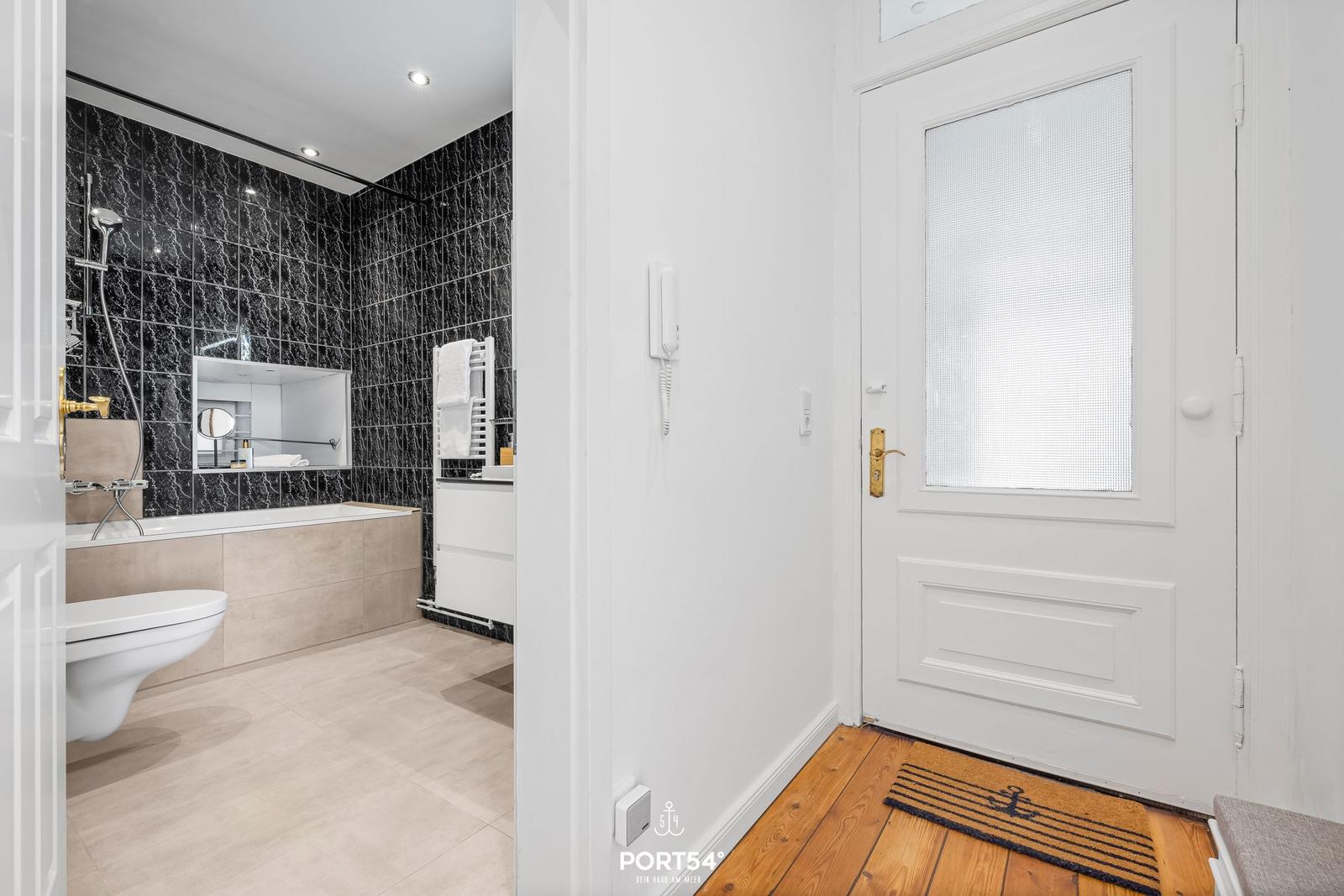 Badezimmer mit schwarzen Fliesen, Badewanne und Toilette. Flur mit Holzboden und weißer Tür.