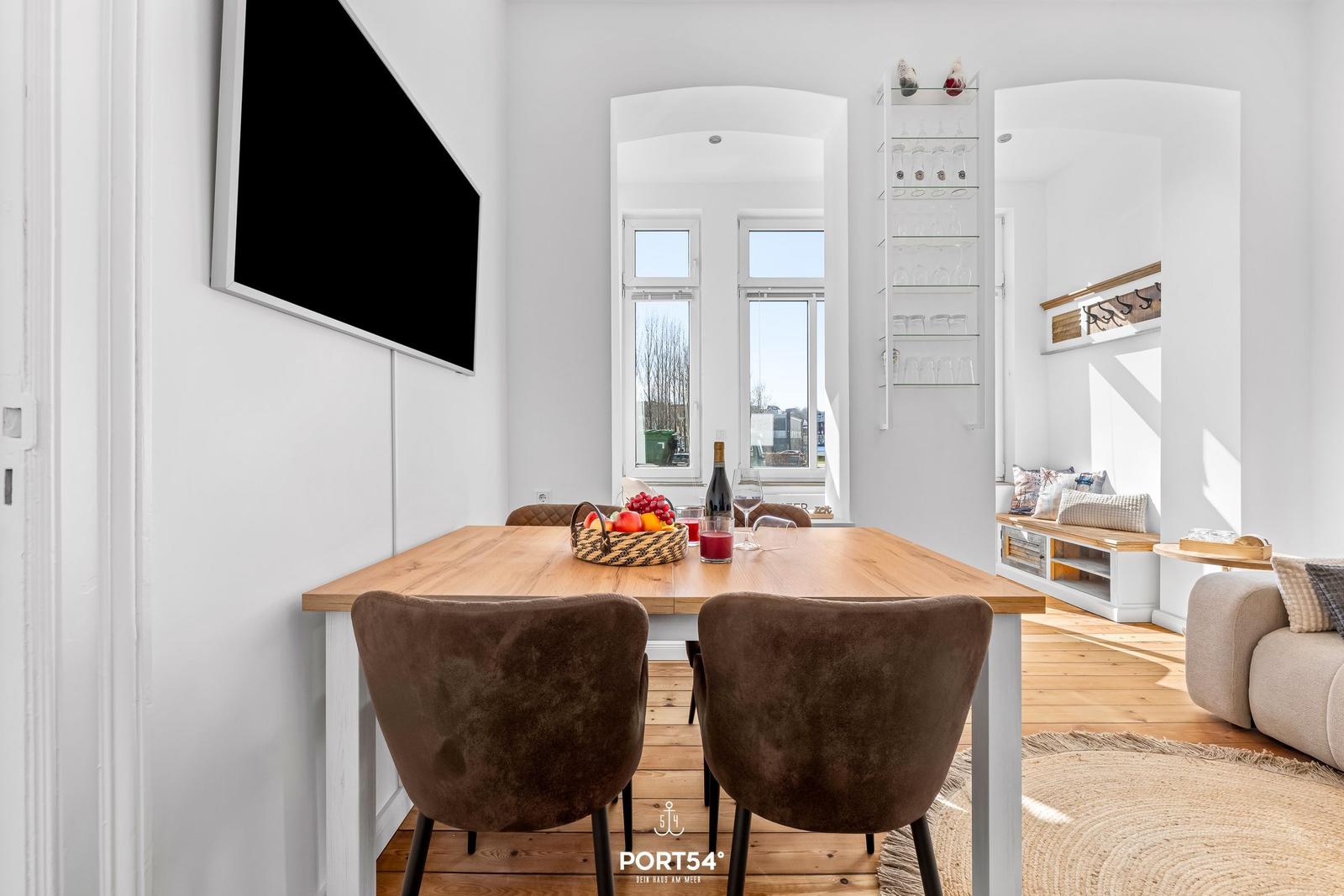 Essbereich mit Holztisch, braunen Stühlen und TV an der Wand. Blick in Wohnzimmer mit Sofa und Fenster.