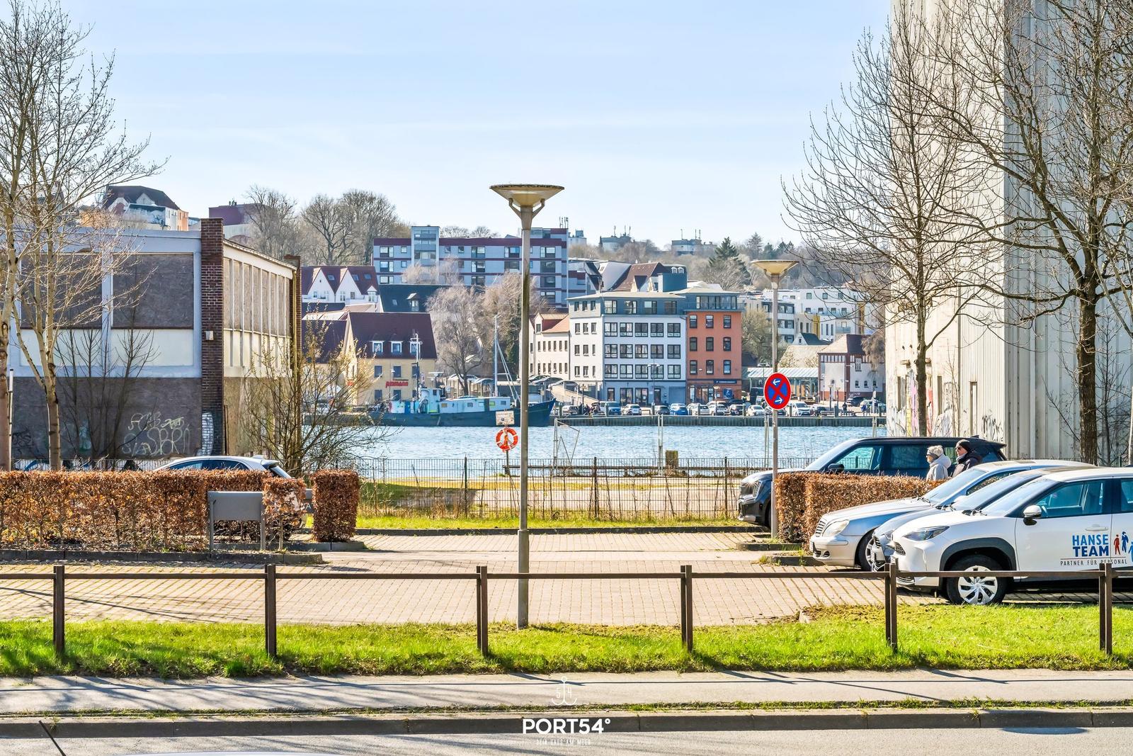 Uferpromenade mit Blick auf Häuser und Wasser. Parkplatz mit Autos und Bäumen.