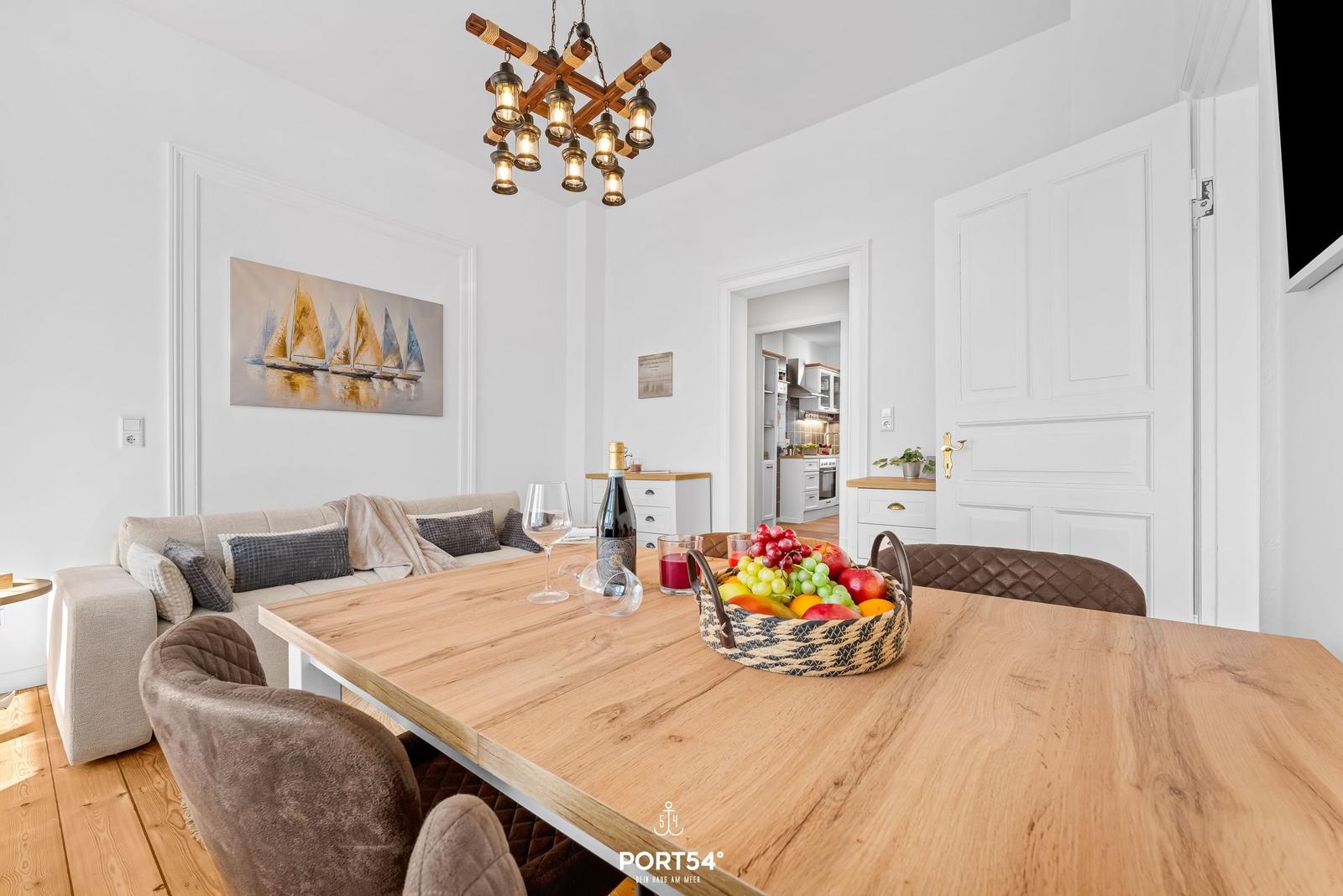 Essbereich mit Holztisch, Sitzgelegenheiten und Obstschale. Küche im Hintergrund.