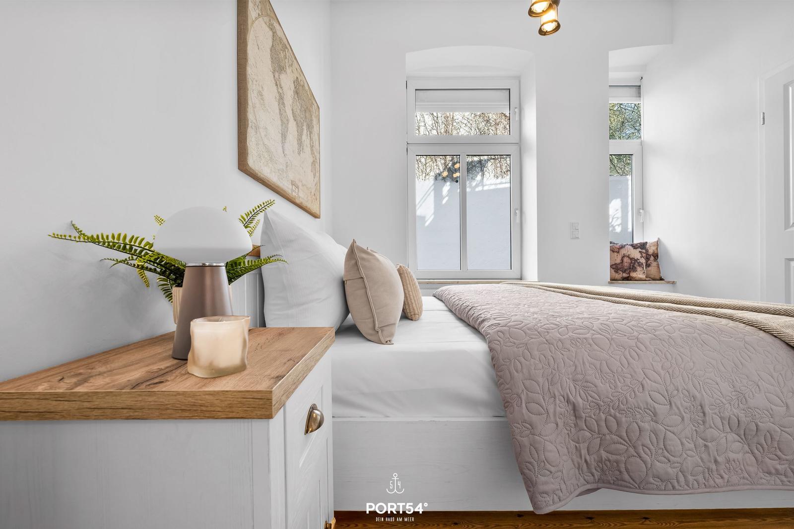 Schlafzimmer mit Bett, Nachttisch und Fenster. Wandbild und Pflanze auf Nachttisch.