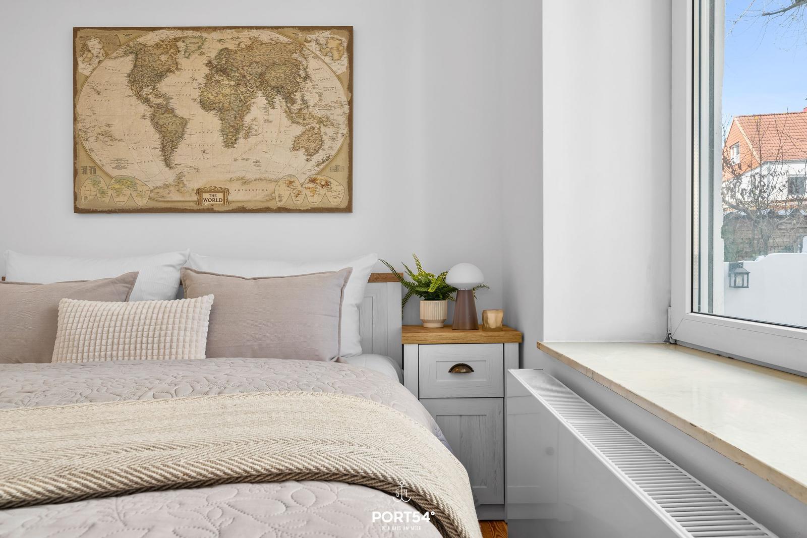 Schlafzimmer mit Bett, Nachttisch und Weltkarte an der Wand. Blick aus dem Fenster.