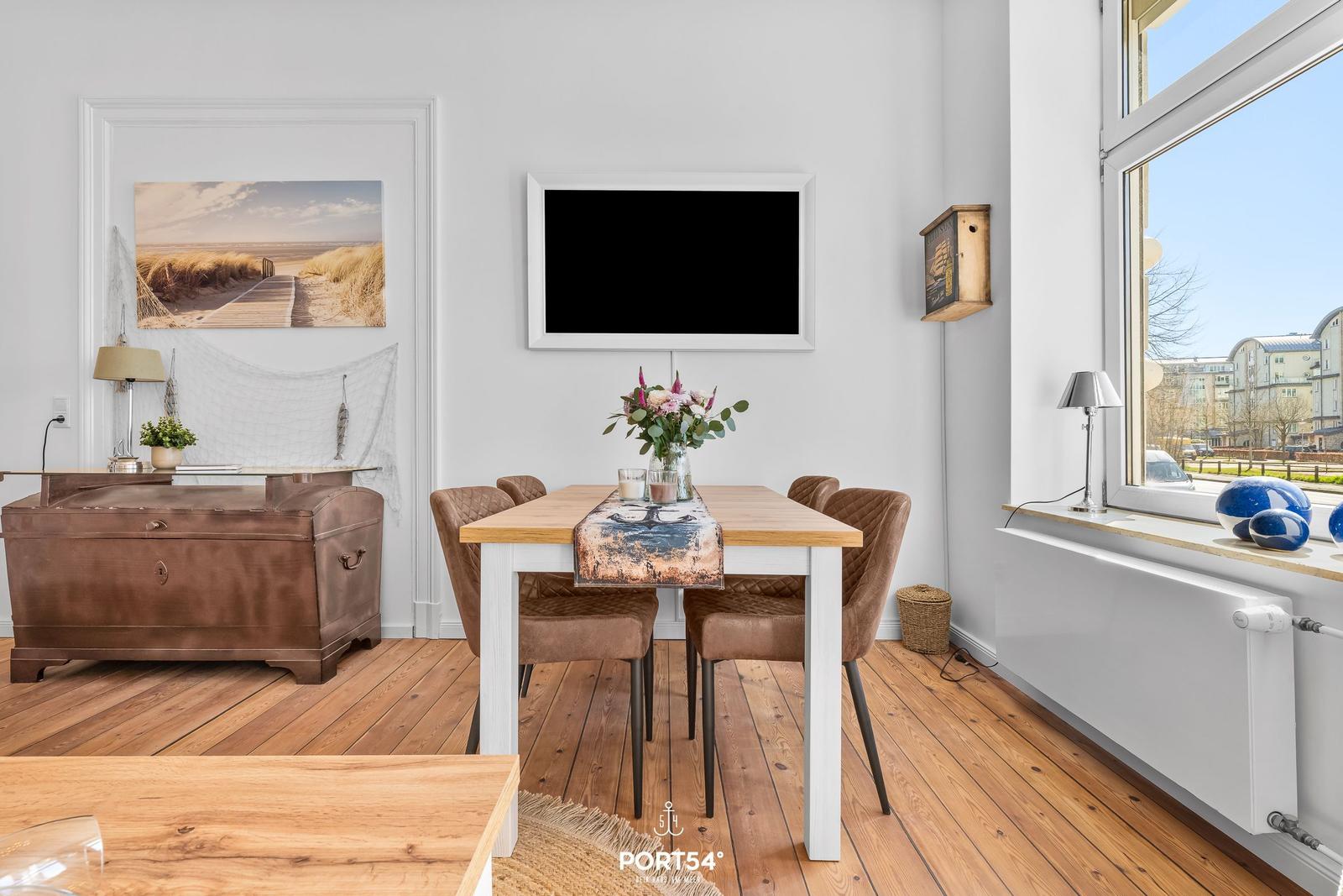 Esszimmer mit Holztisch, Stühlen und Fensterblick. Wandbild und Fernseher an der Wand.