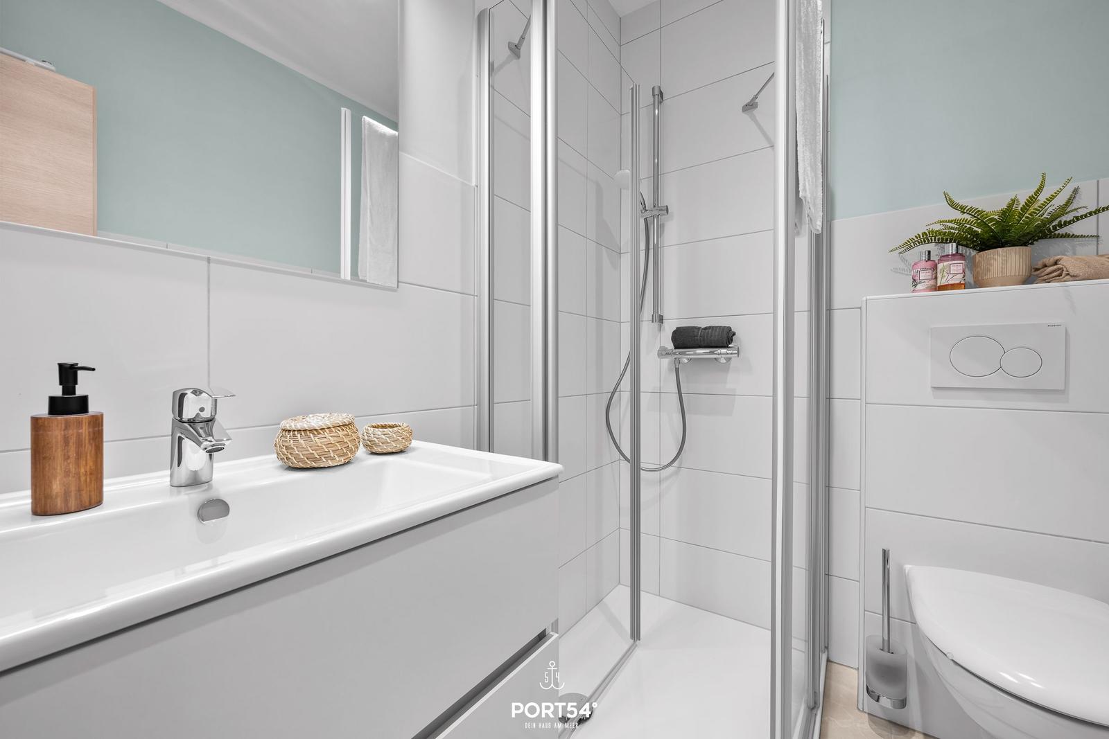 Badezimmer mit Dusche, Waschbecken und Toilette. Weiße Fliesen und moderne Ausstattung.