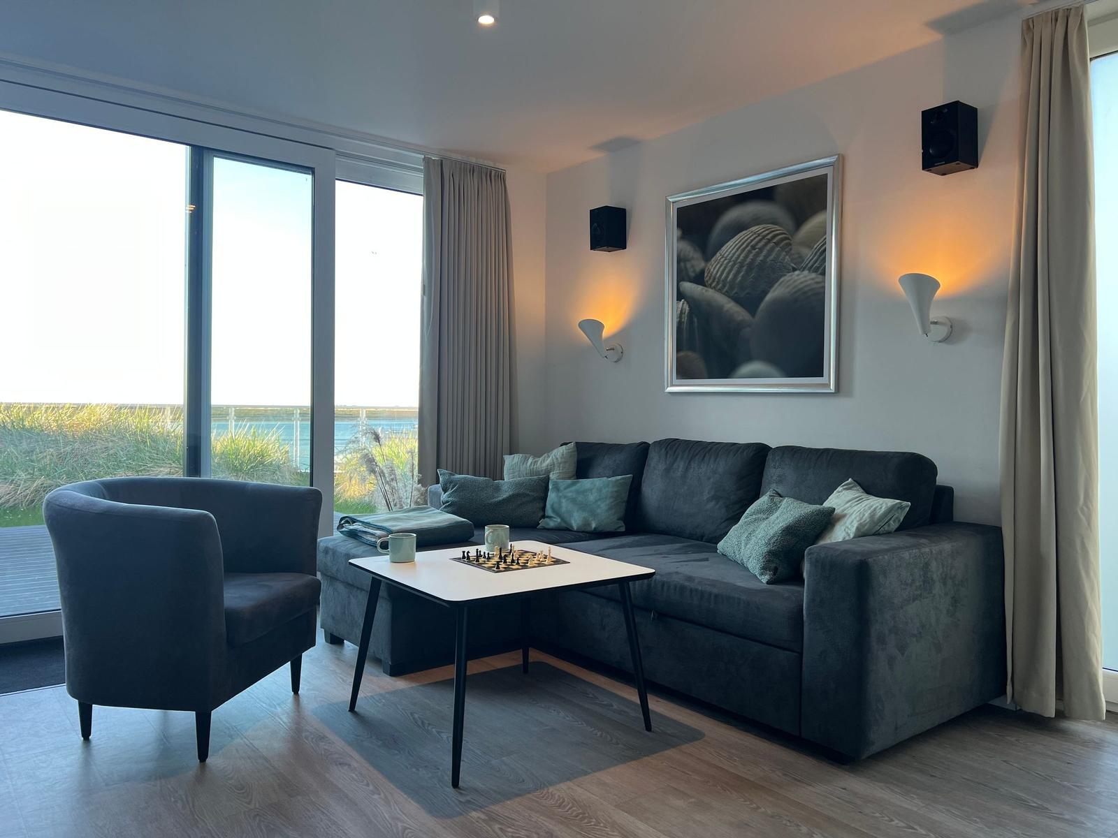 Wohnzimmer mit Sofa, Sessel und Tisch. Blick auf See und Strand.