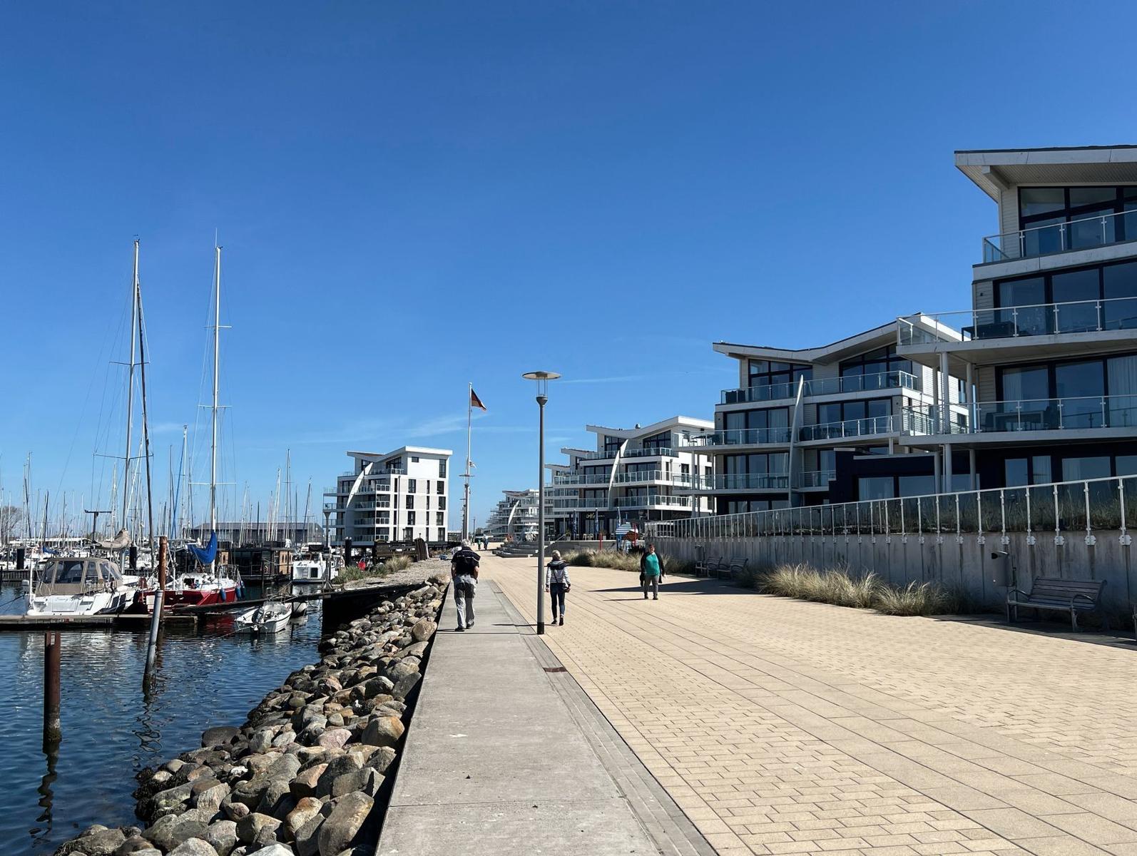 Promenade am Hafen mit modernen Gebäuden und Anlegestellen für Segelboote.
