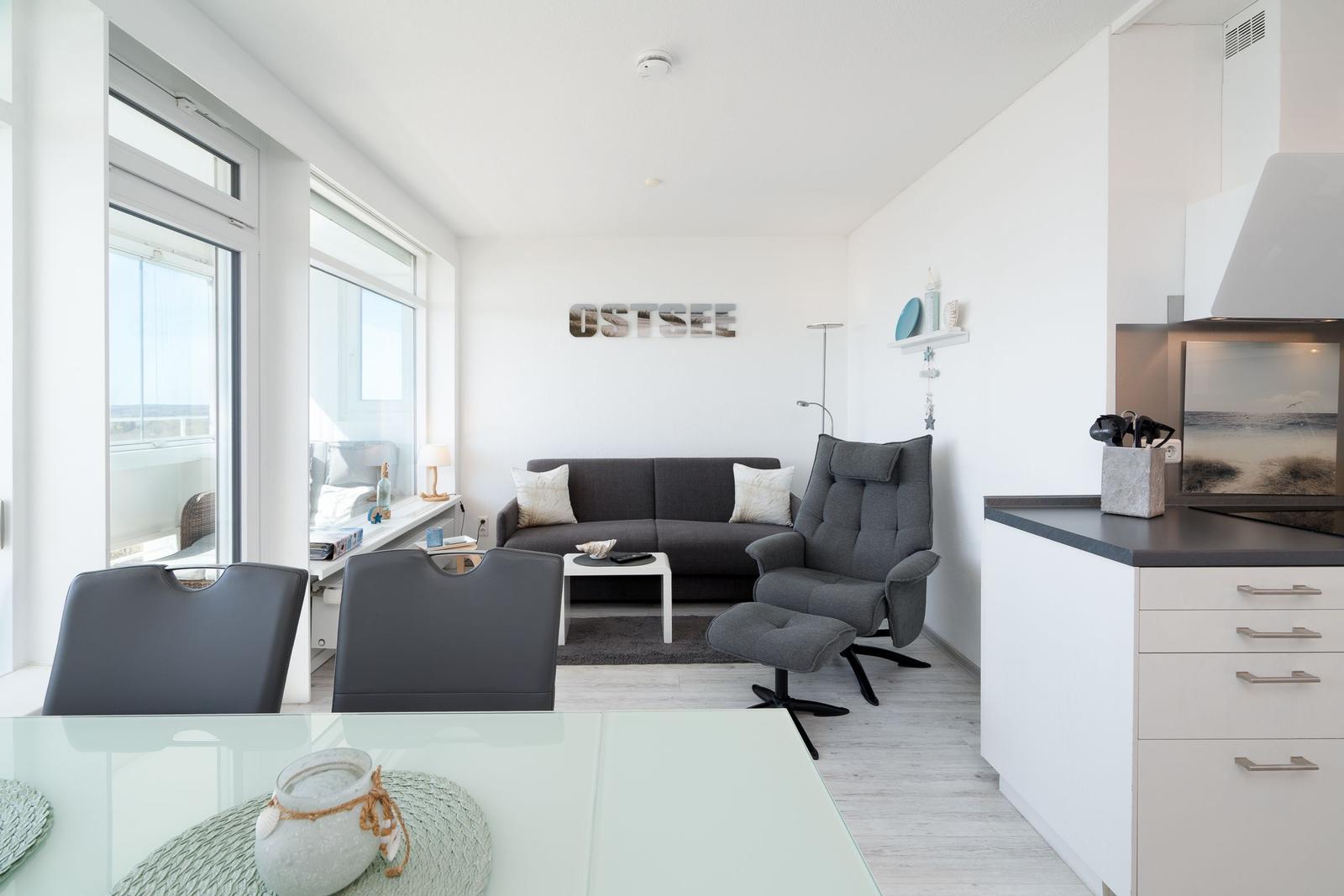 Living area with sofa, armchair, and kitchen. Large windows with sea view.