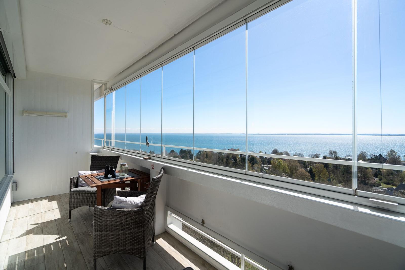 Balcony with table and chairs overlooking the sea through large glass windows.