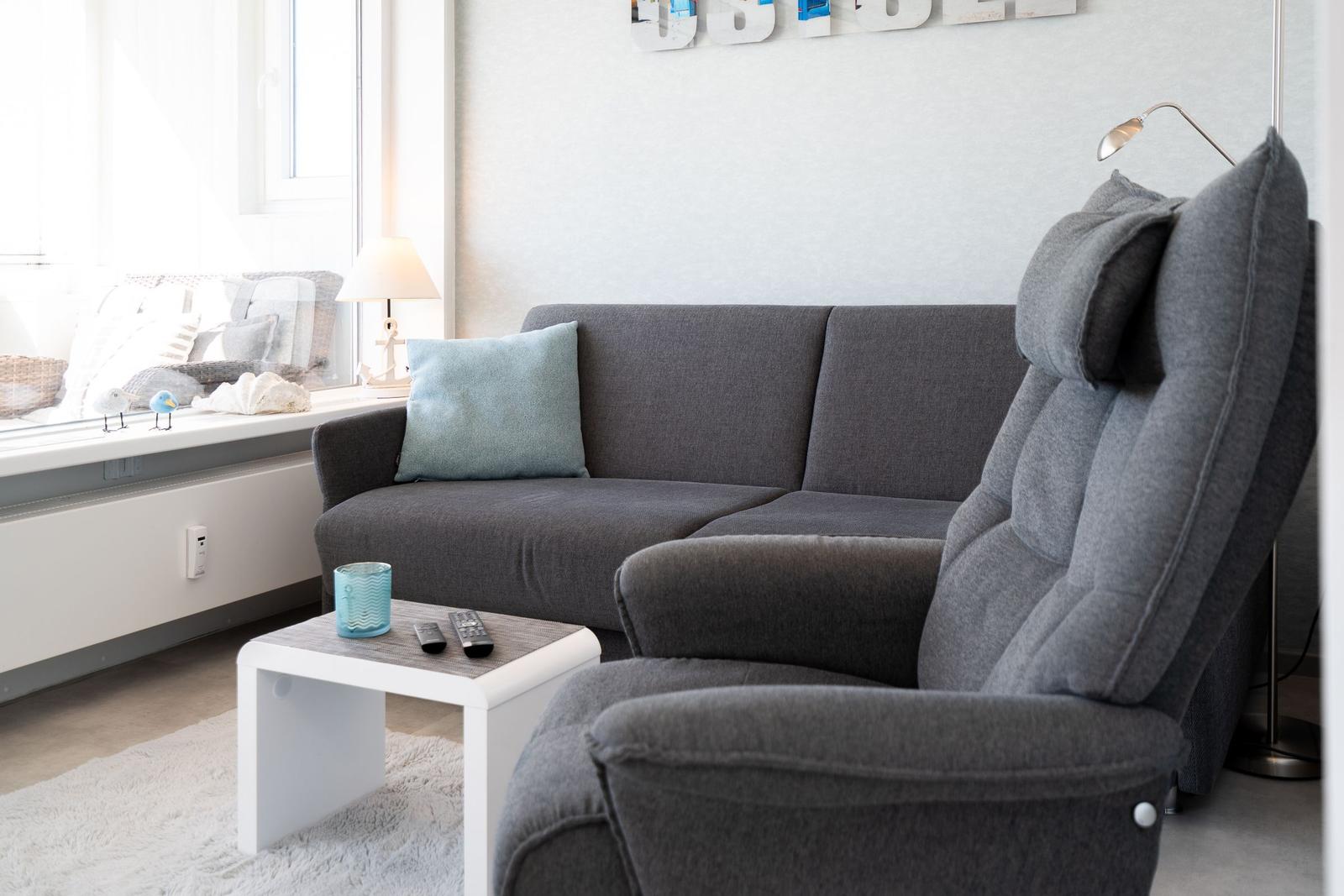 Ein grauer Sofa und Sessel stehen vor einem Fenster mit Tisch und Kissen.