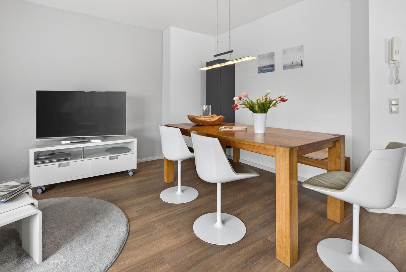 Living and dining area with wooden table, white chairs, TV, and flowers.