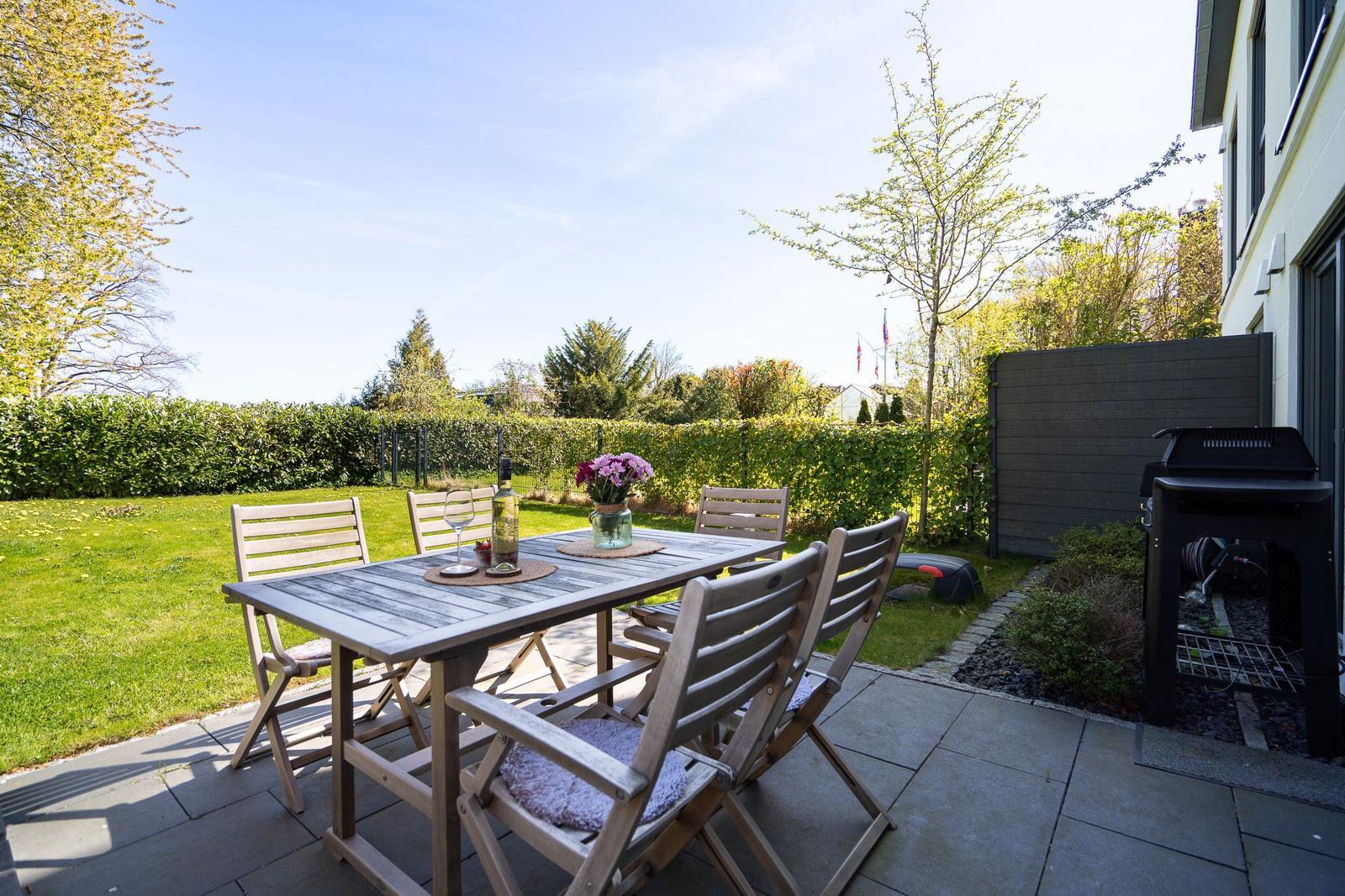 Terrace with table, chairs, and grill. Lawn and hedge in the background.