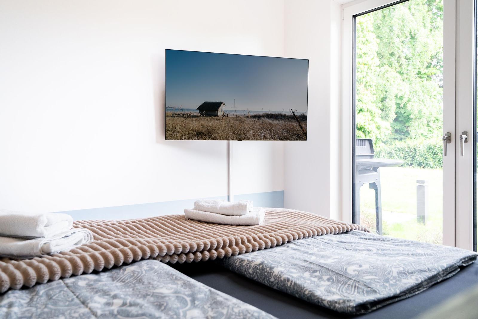 Bedroom with bed, towels, and TV on wall. Doorway to terrace with view of green areas.