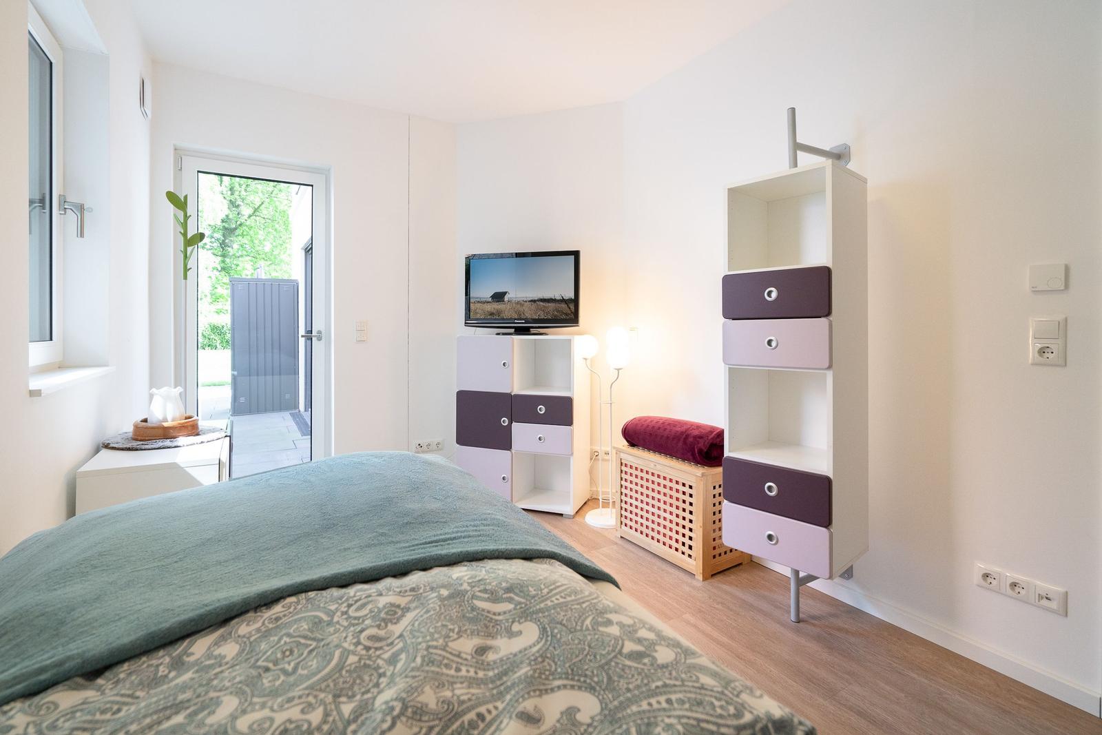Bedroom with bed, TV, and cabinets. Door leads to balcony. Wooden floor and white walls.