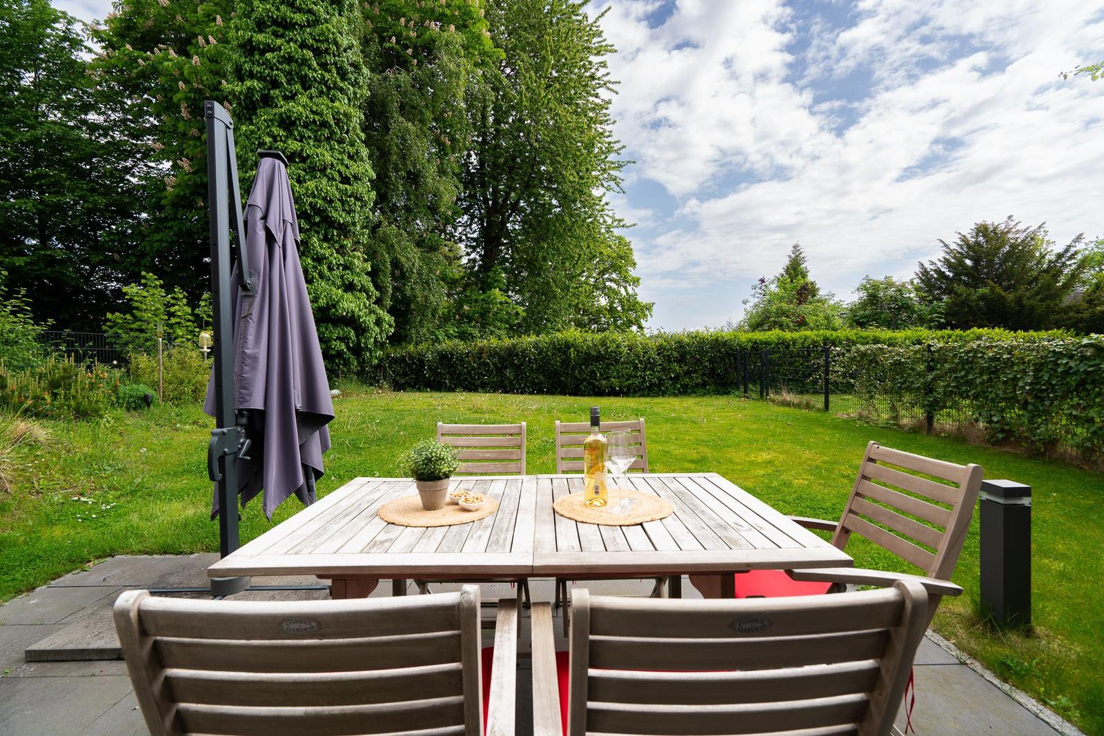 Terrace with table, chairs, and sunshade. Lawn and hedge in the background.