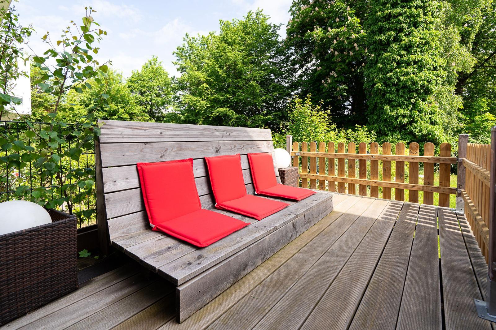 Wooden terrace with red cushions and wooden railing surrounded by greenery.