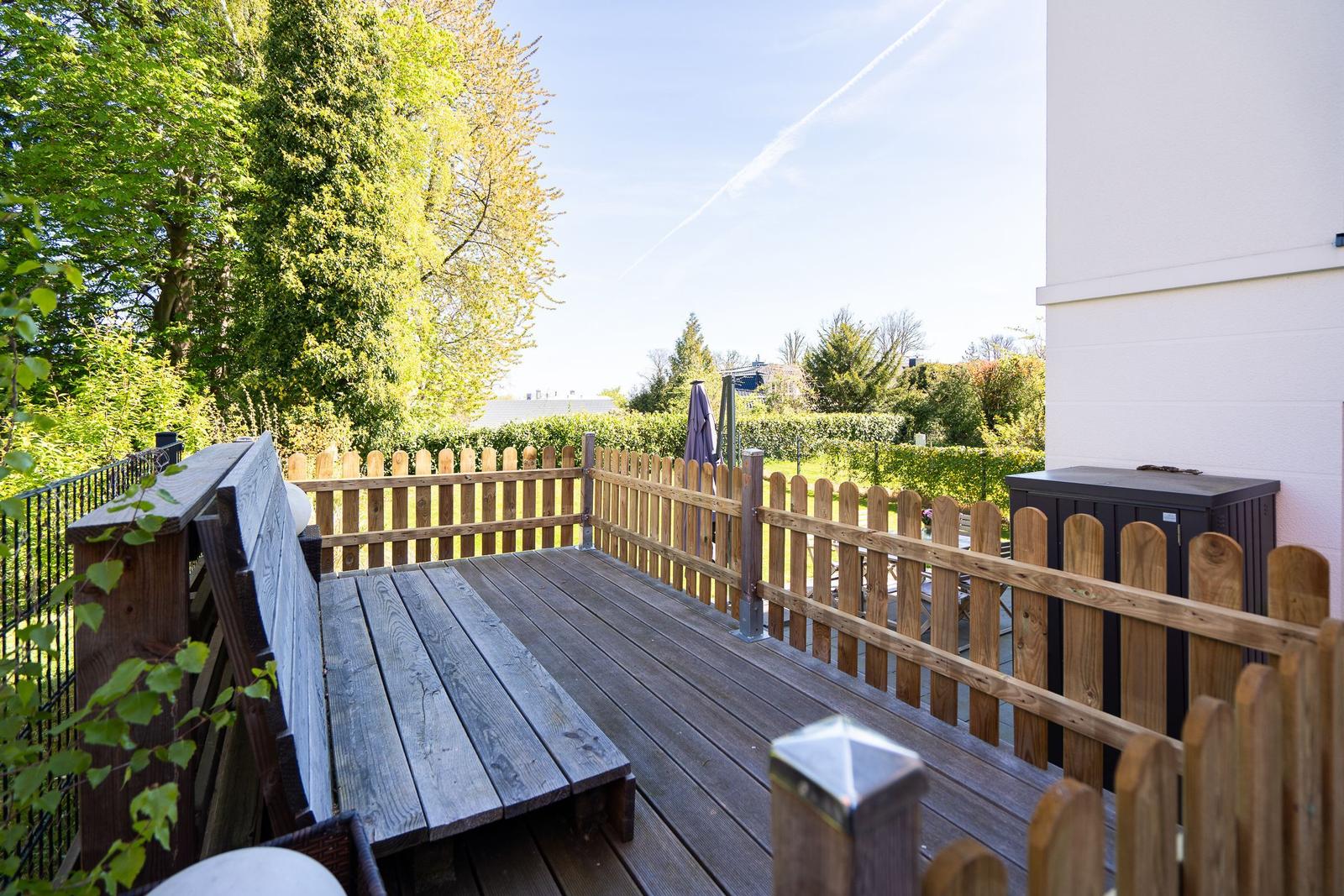 Wooden terrace with benches and fence, view of green garden and sky.