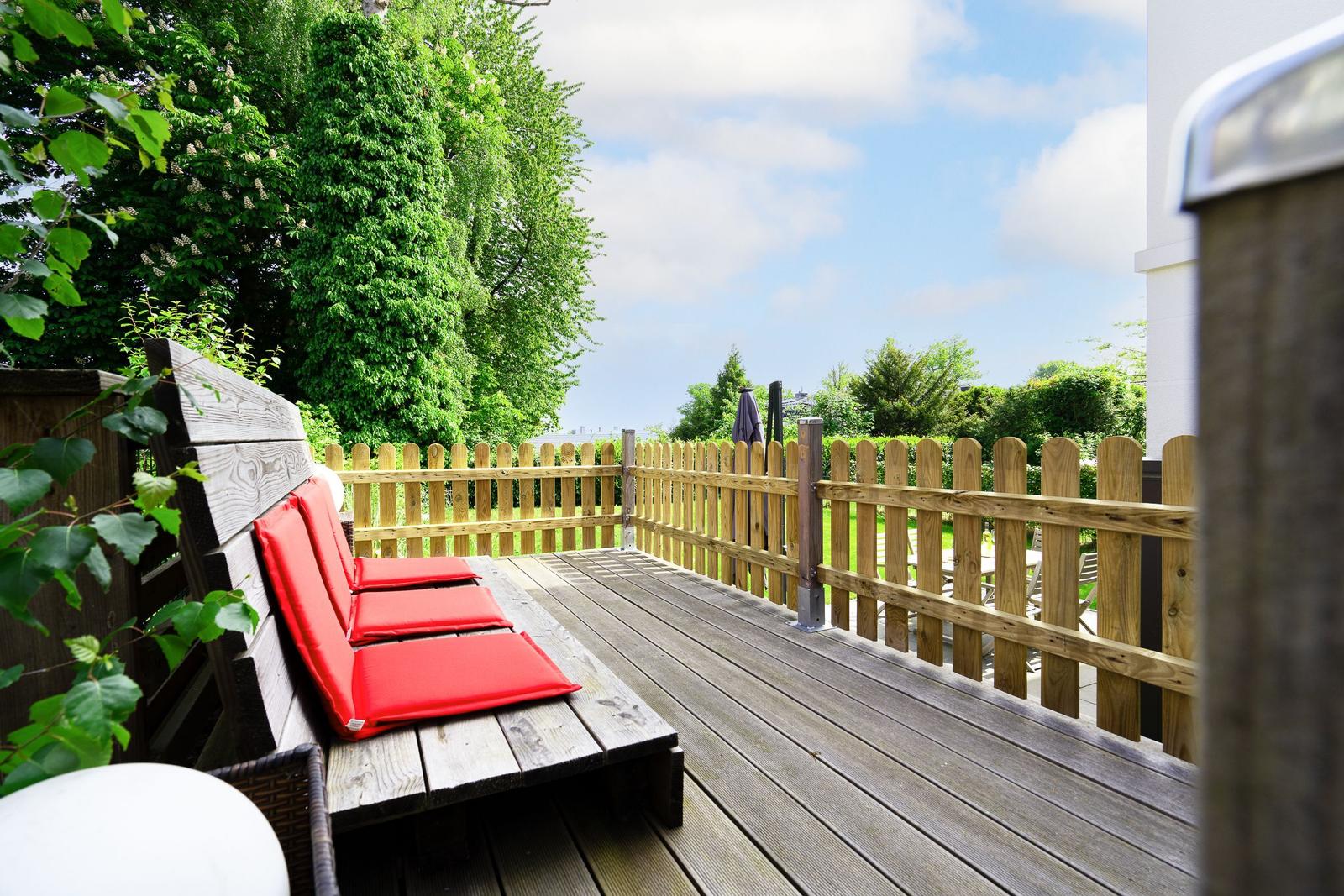 Wooden terrace with red bench and fence. Green trees and sky in the background.