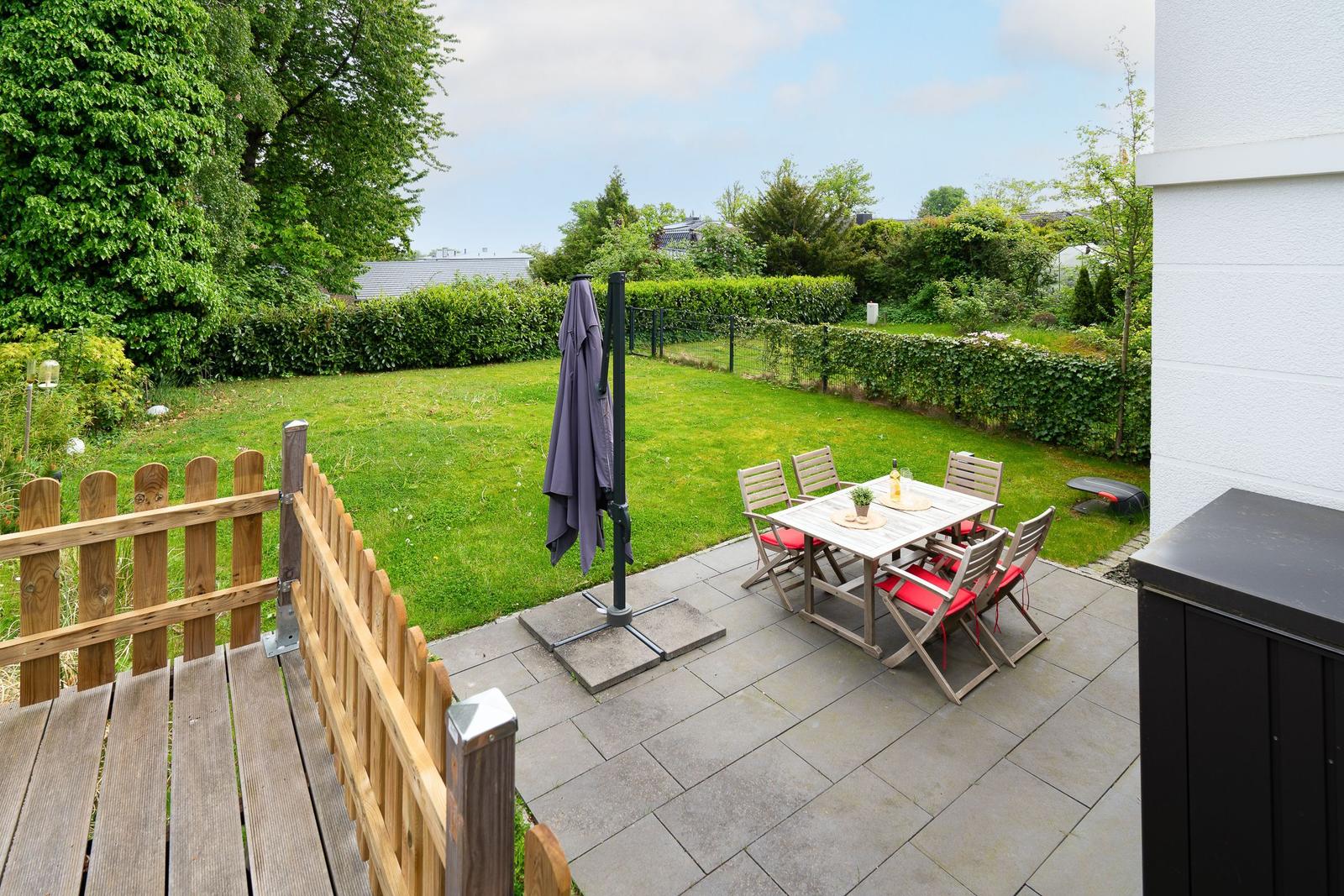 Terrace with table, chairs, and umbrella. Backyard with lawn and hedge.