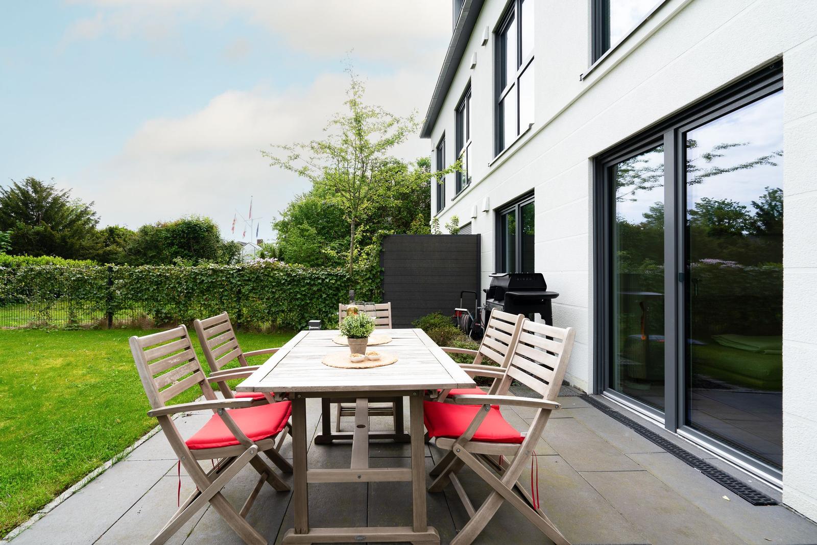 Deck with wooden table, red seat cushions, and grill beside the house.
