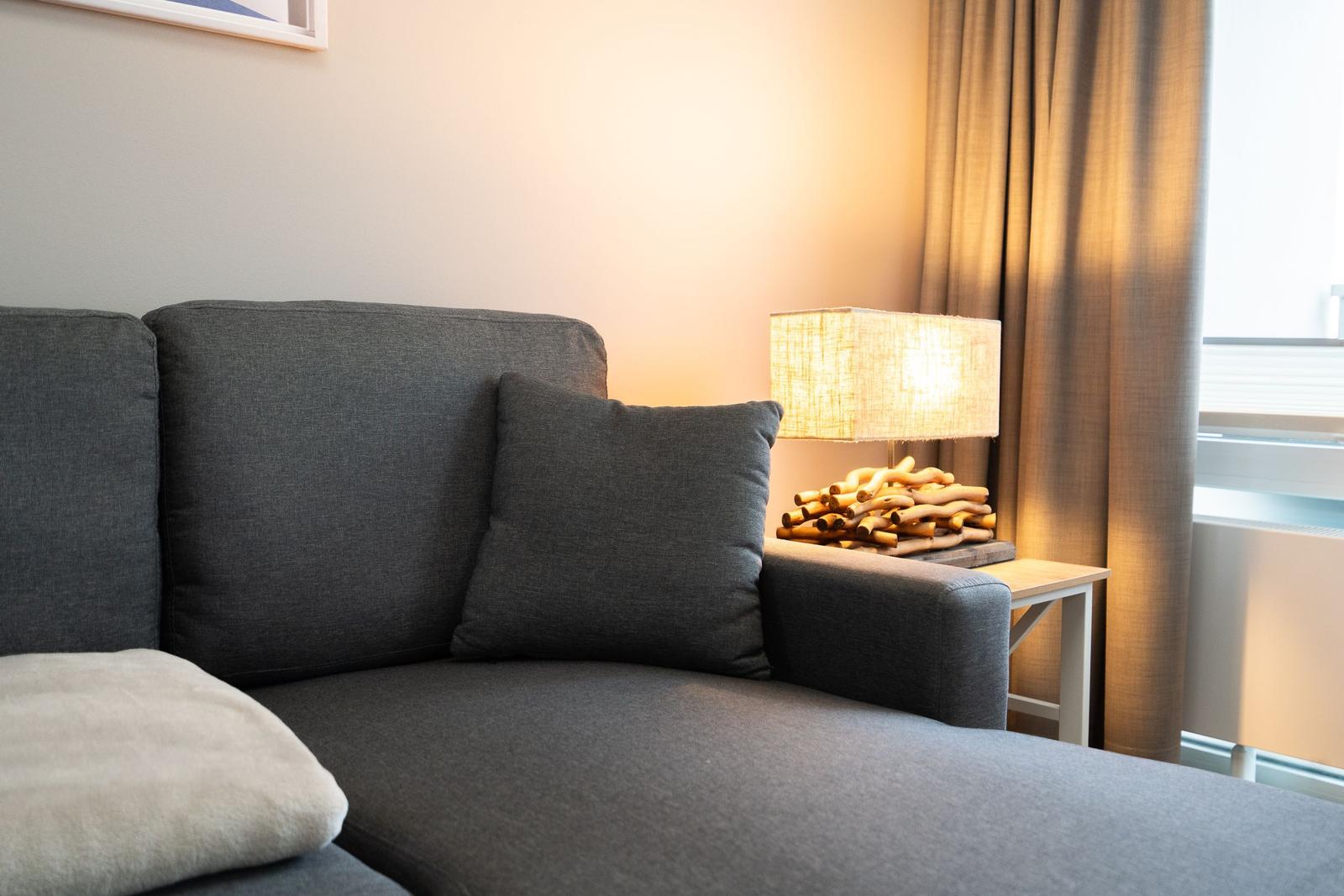 A gray sofa with cushions and a lamp on a table next to a window.