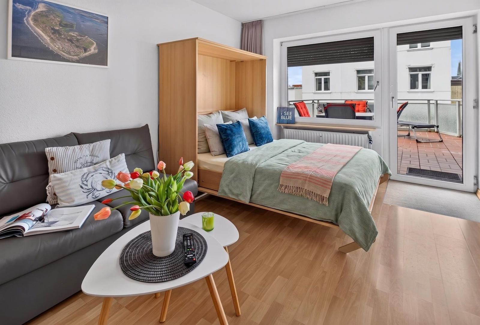 Wohnraum mit Schlafcouch, Tisch und Blumen. Balkon mit Sitzgelegenheiten.