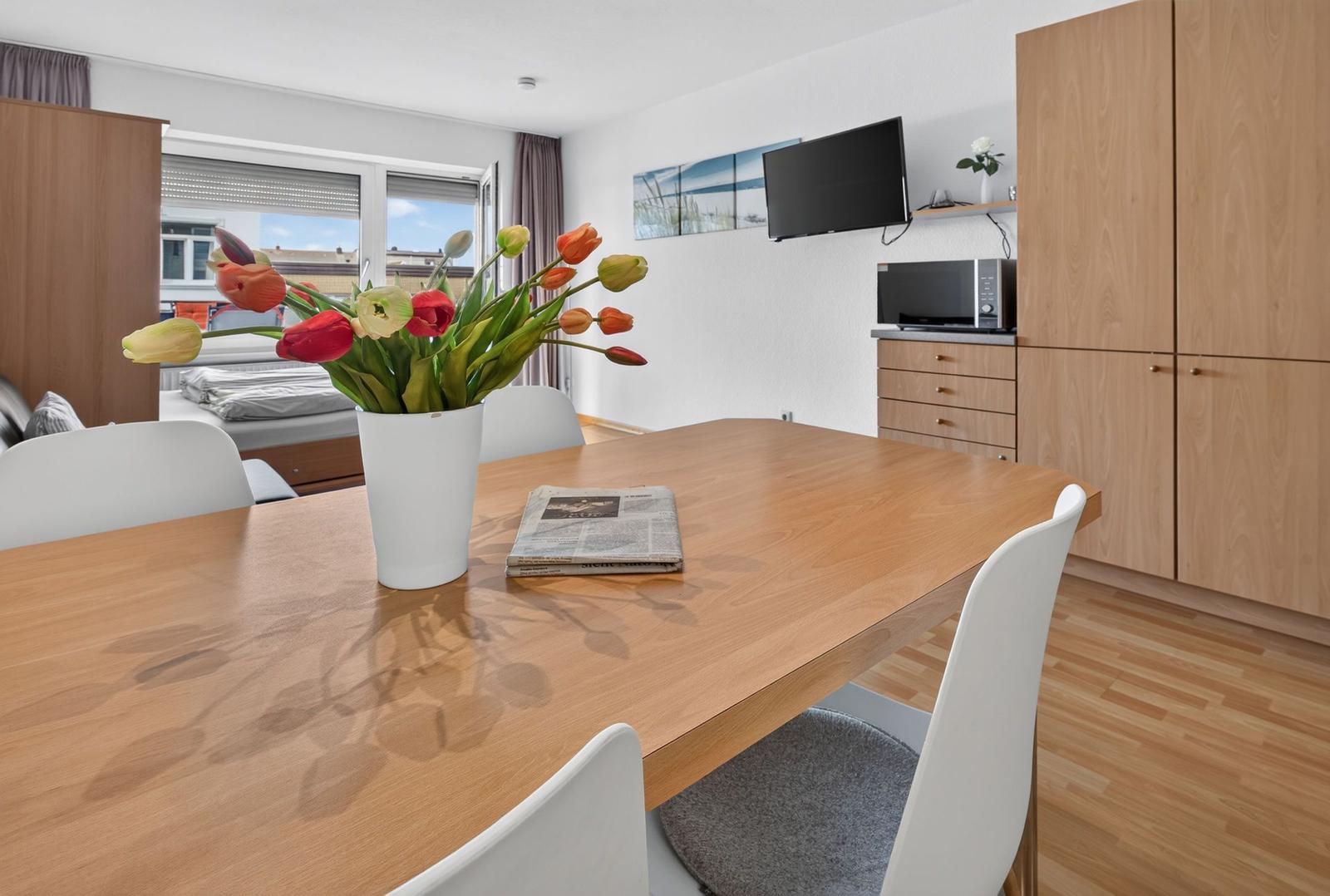 Essbereich mit Holztisch, weißen Stühlen und Blumen. Im Hintergrund Küche und Schlafbereich.