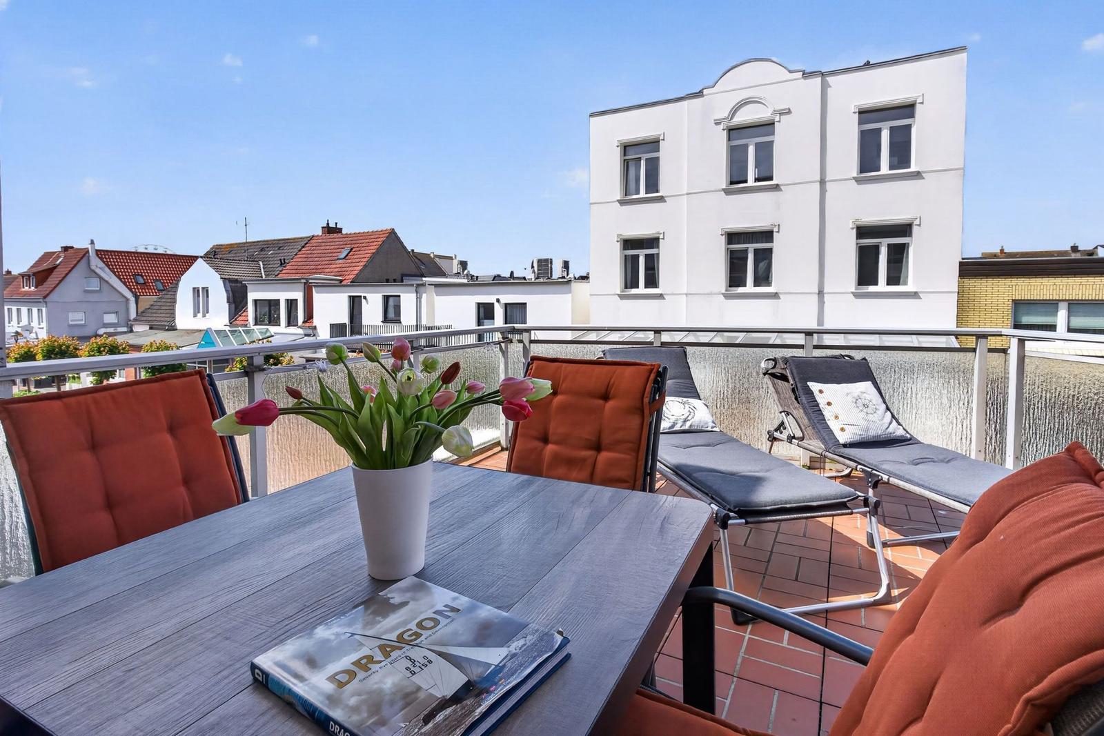 Terrasse mit Tisch, Stühlen und Blumen. Blick auf Wohnhäuser und Himmel.