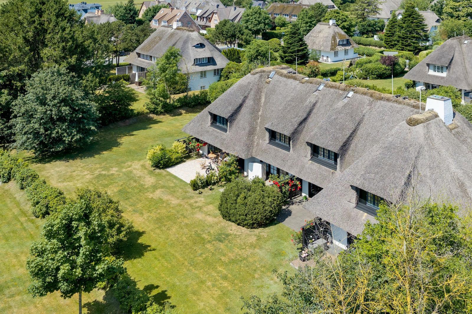 Dorfhaus mit Strohdach, großem Garten und Terrasse. Umgeben von Bäumen und weiteren Häusern.
