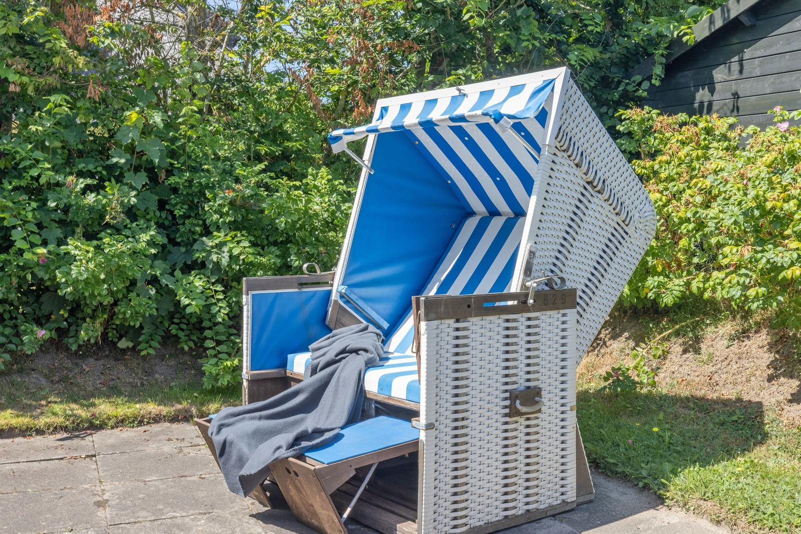 Weiße Strandkorb mit blau-weißem Stoff und dunkler Decke im Garten.