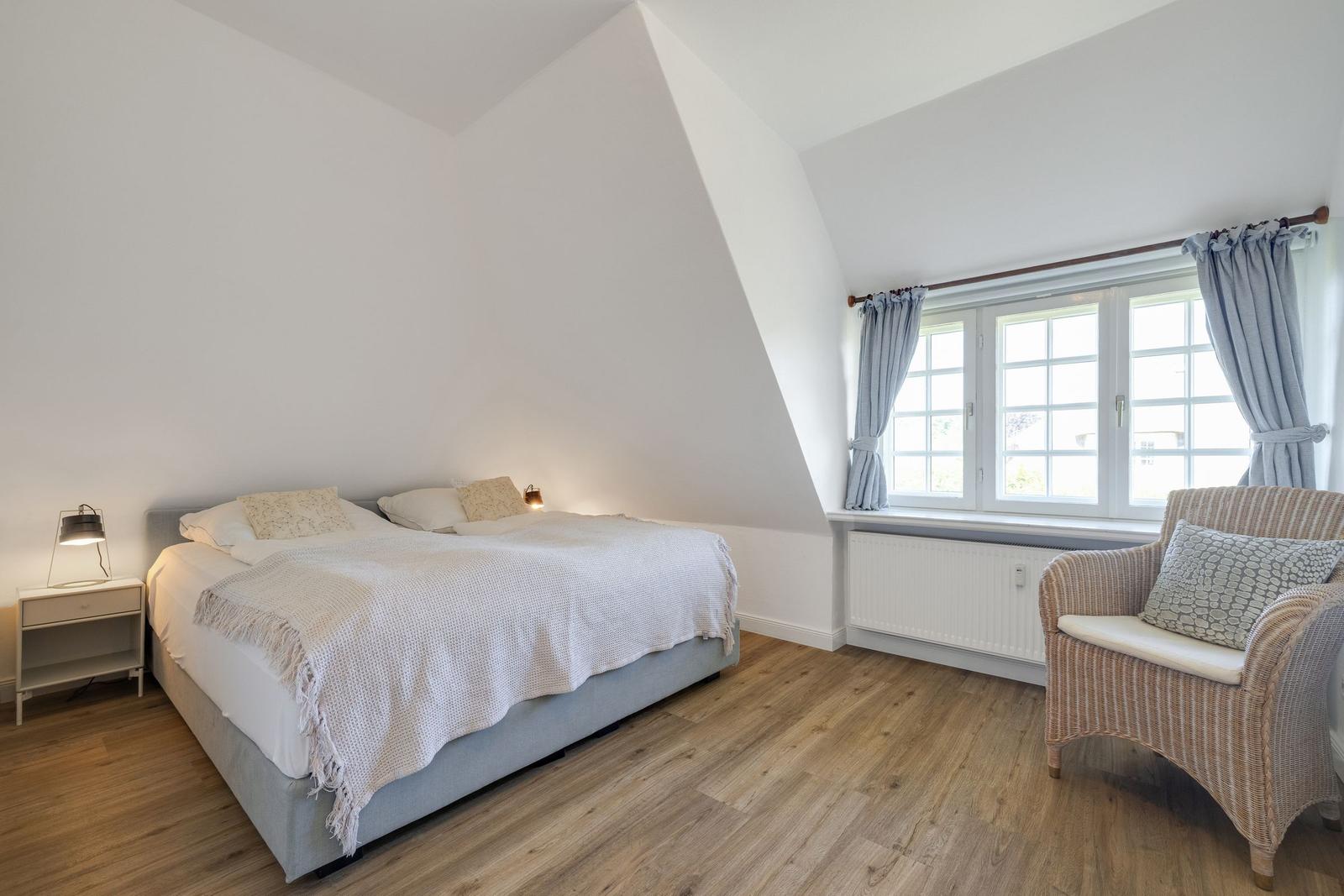 Schlafzimmer mit Bett, Nachttisch und Stuhl neben Fenster mit Vorhängen.