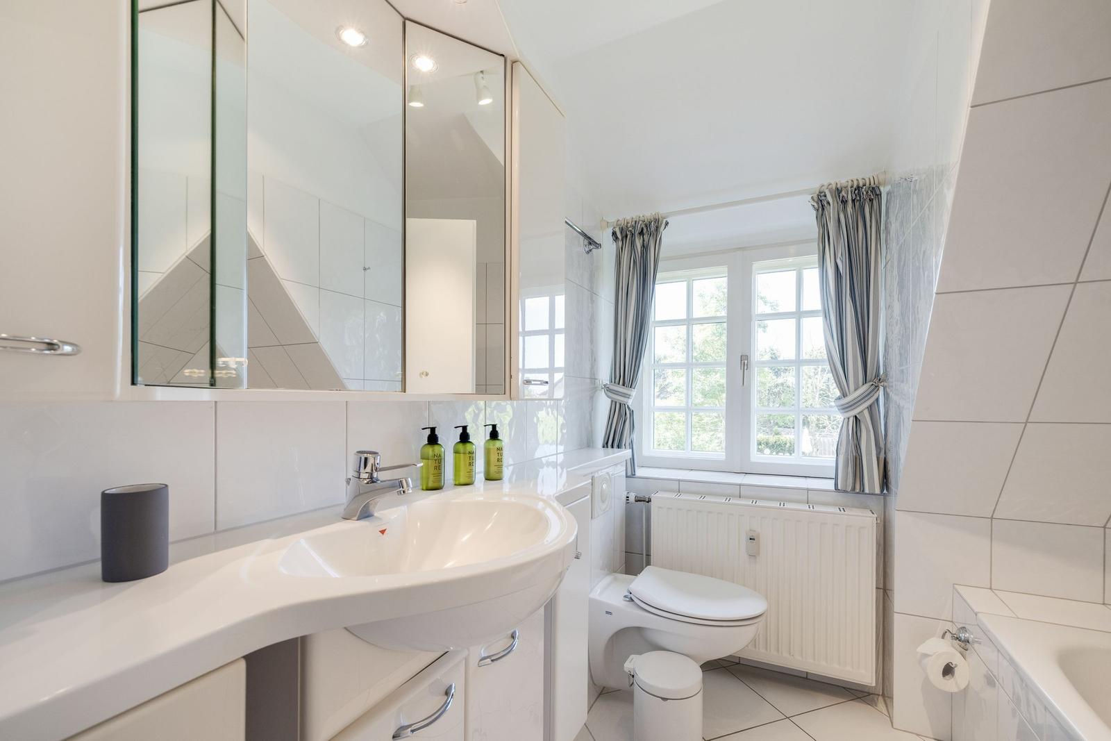 Badezimmer mit Waschbecken, Spiegel, Toilette und Fenster mit Vorhängen.