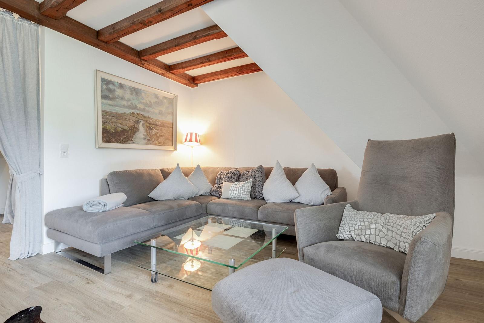 Wohnzimmer mit grauem Sofa, Sessel und Glas-Tisch unter Holzbalkendecke.