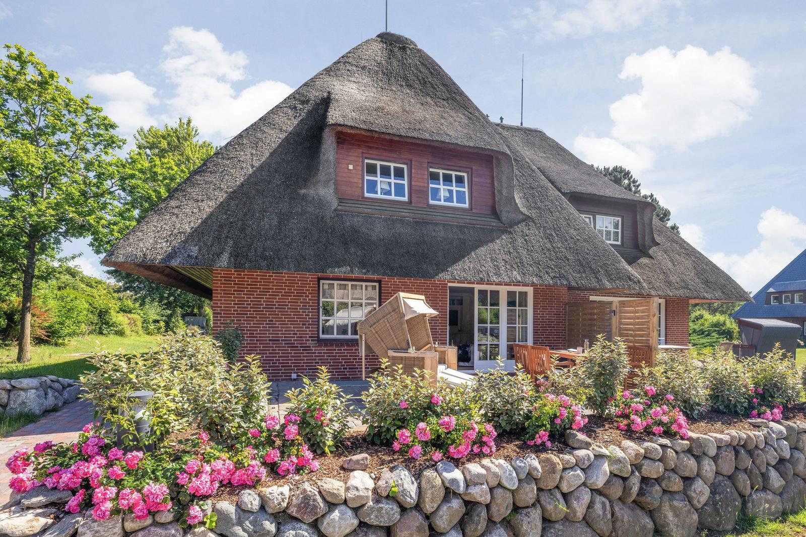 Haus mit Strohdach, Blumenbeet und Steinmauer im Vorgarten.