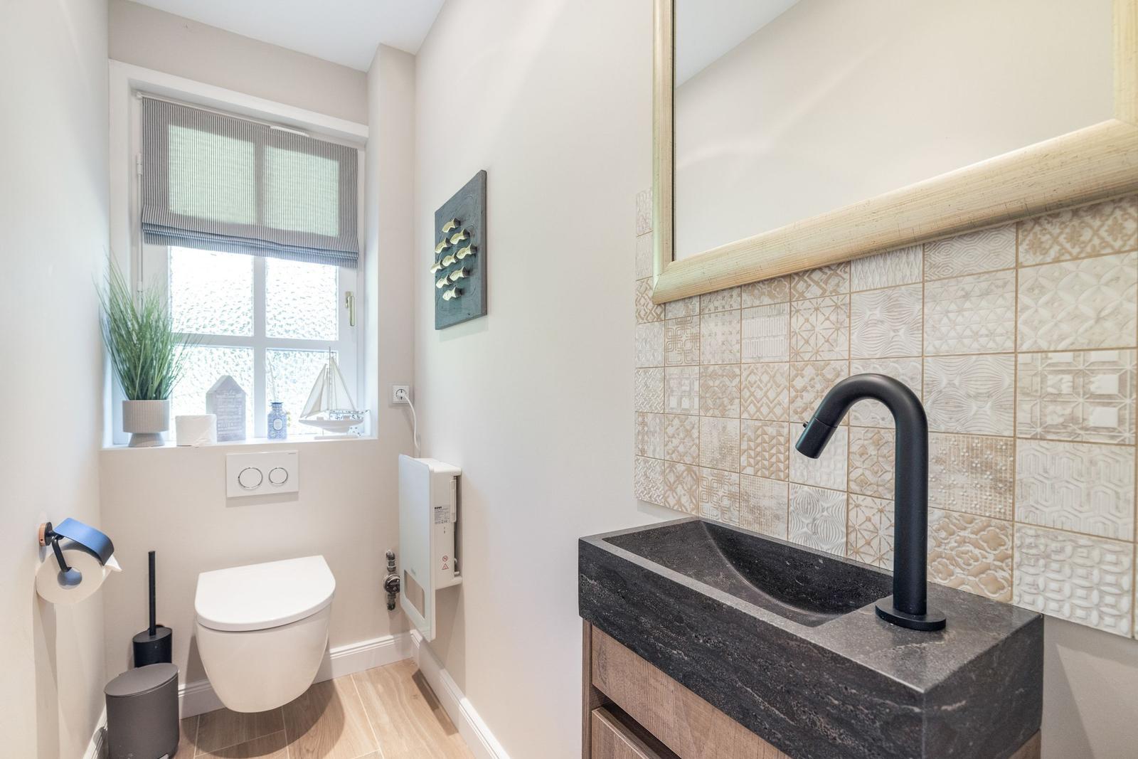 Badezimmer mit Waschbecken, Spiegel und Toilette. Fenster mit Jalousie und Pflanze.