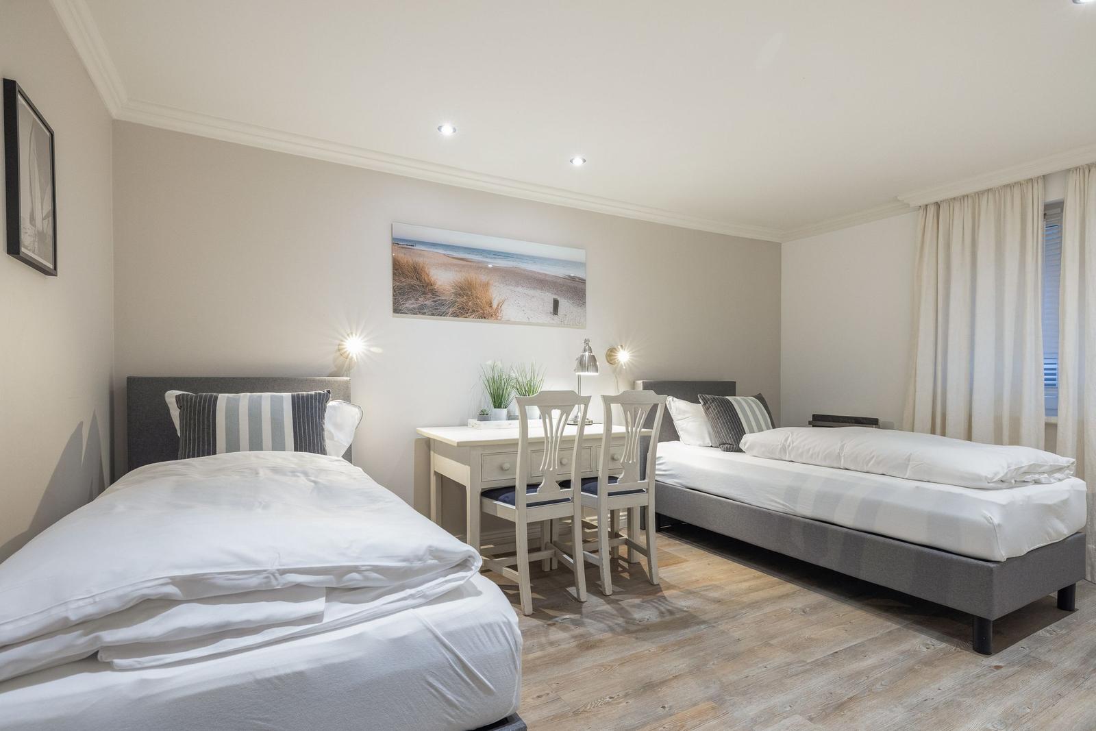Zwei Betten, Schreibtisch mit Stühlen und Wandbild mit Strandmotiv.